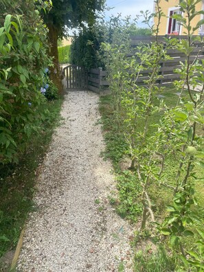 Un chemin de gravier mène à un portail en bois, entouré de verdure et bordé d’un bâtiment à la façade jaune.