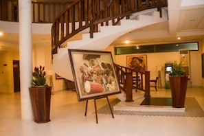 A hotel lobby with a wooden staircase, a framed painting on an easel, and potted plants. - Hôtel Sancta Maria  (Lomé)