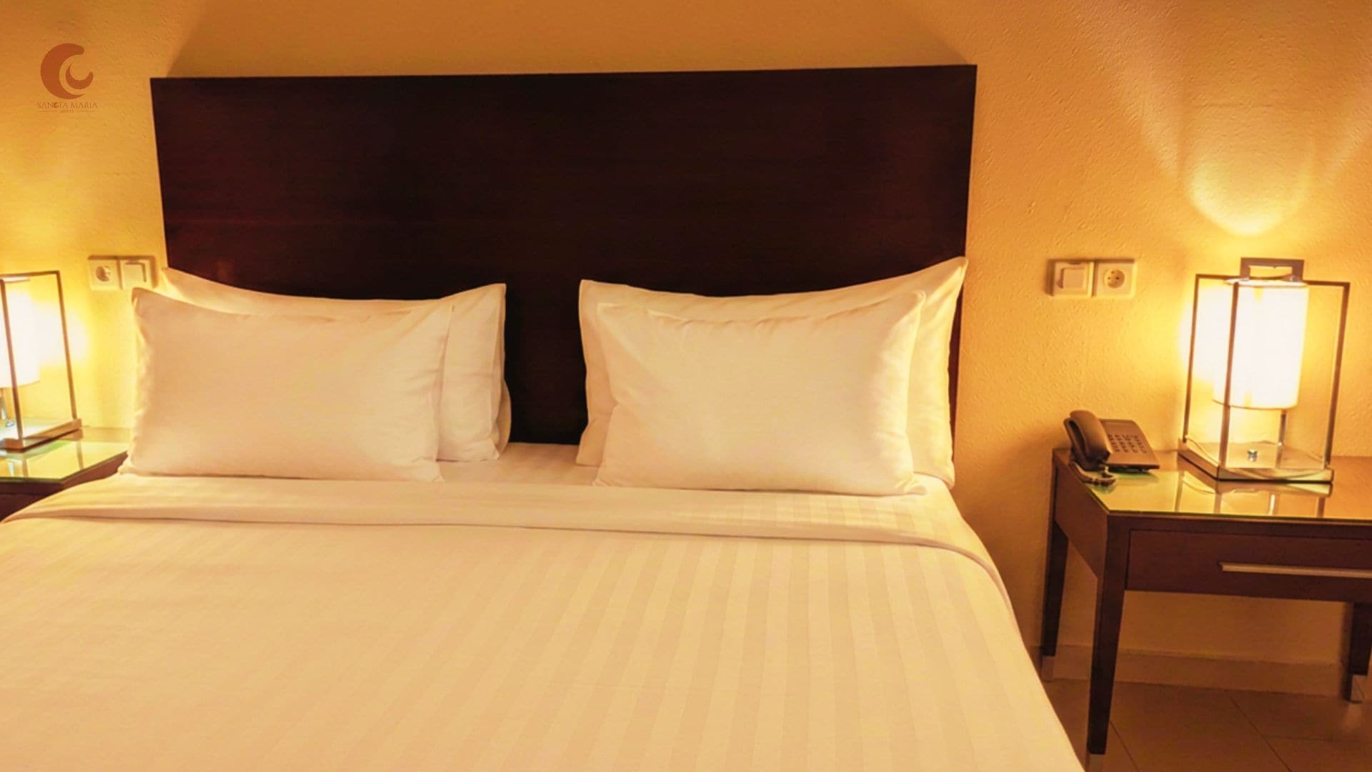 A hotel room with a large bed, two bedside lamps, a nightstand with a phone, and a dark headboard.