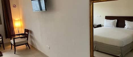 Una habitación de hotel con cama, televisión montada en la pared, una silla y una lámpara.
