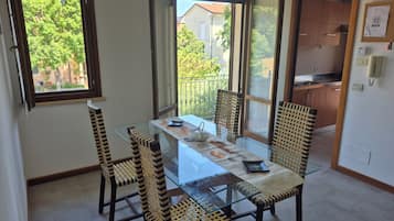 Spiseområde med et glasbord og stole, udsigt til det fri og et køkken i baggrunden.