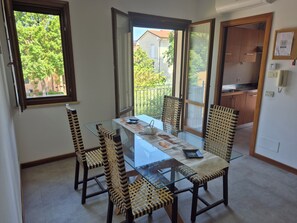 A dining area with a glass table and chairs, a view of the outside, and a kitchen in the background.