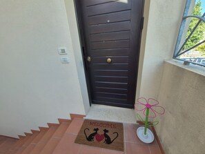 A dark wooden door with horizontal panels and a round handle, a doormat with a cat and heart design, and a small potted plant with a pink flower.