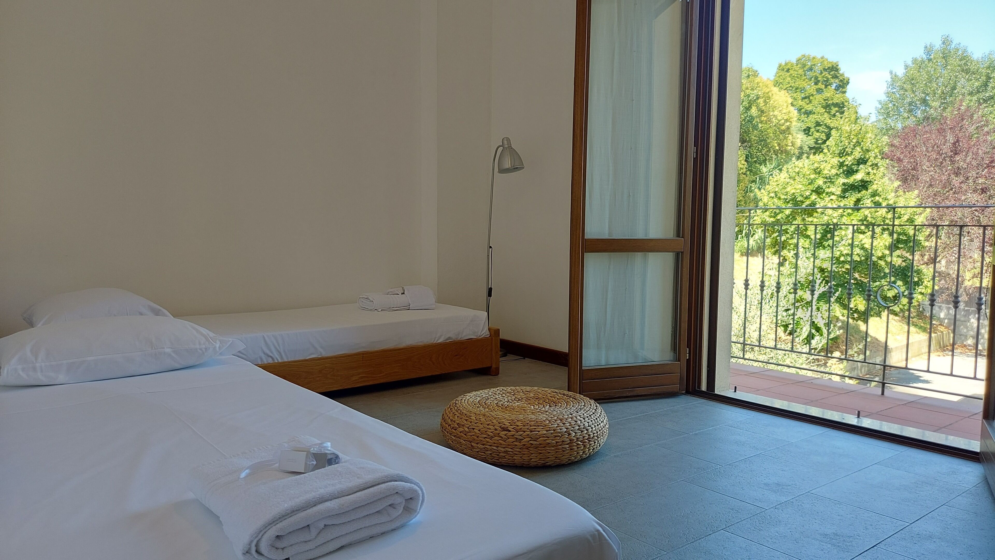 Hotelkamer met een bed, een bedlamp, een klein rond rieten pouf en uitzicht op een balkon met een hekwerk.