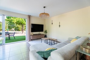 Ett modernt vardagsrum med en stor TV, en soffa, en matplats med bord och stolar samt utsikt över grönområden utanför.