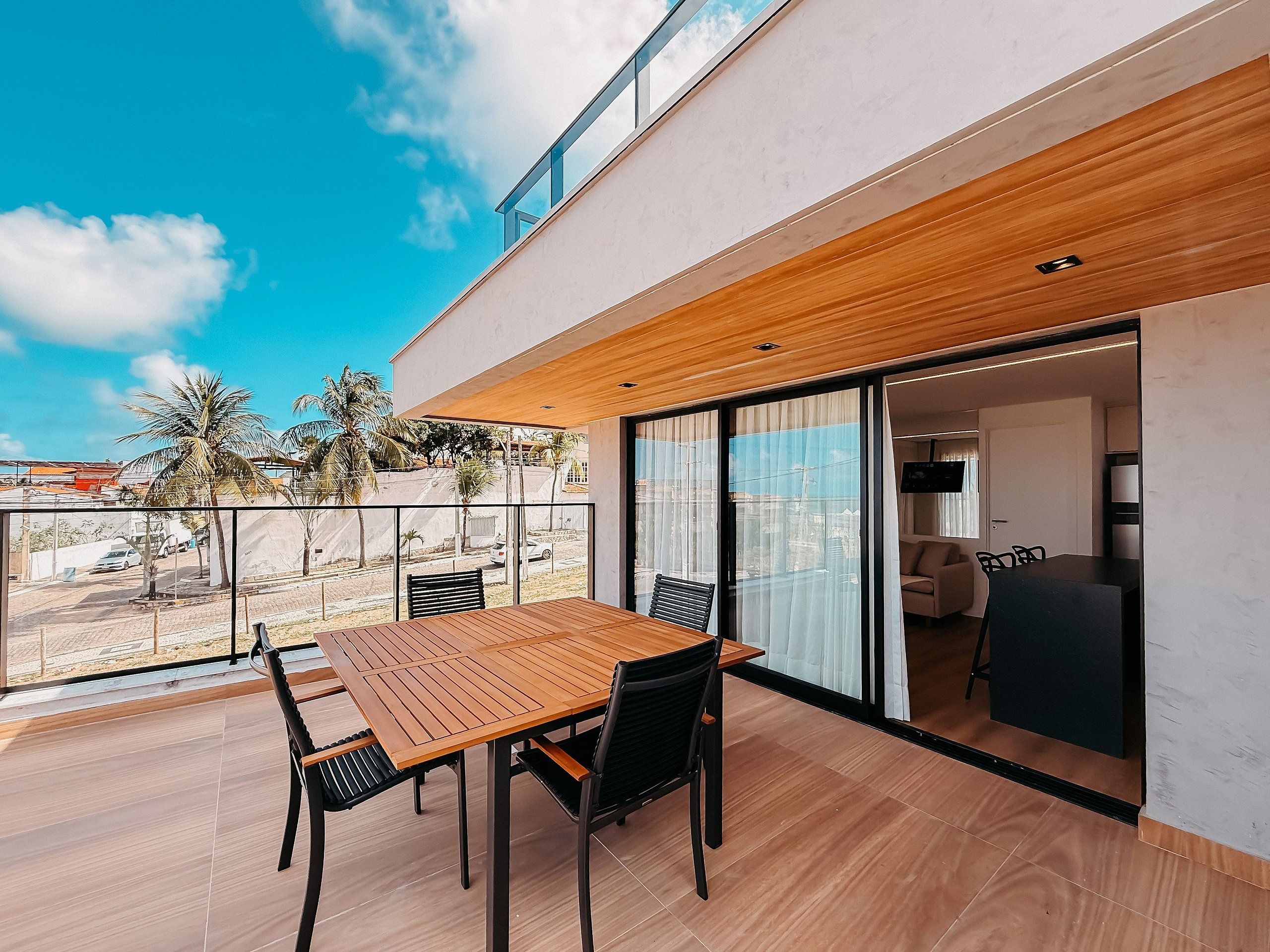 A modern outdoor patio with a wooden table and chairs, a glass door leading to an interior space, and a view of palm trees and a street.