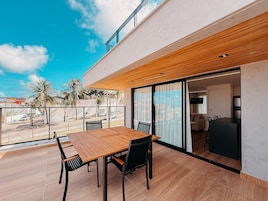 Um pátio moderno ao ar livre com mesa e cadeiras de madeira, porta de vidro que dá para um espaço interior e vista para palmeiras e uma rua.