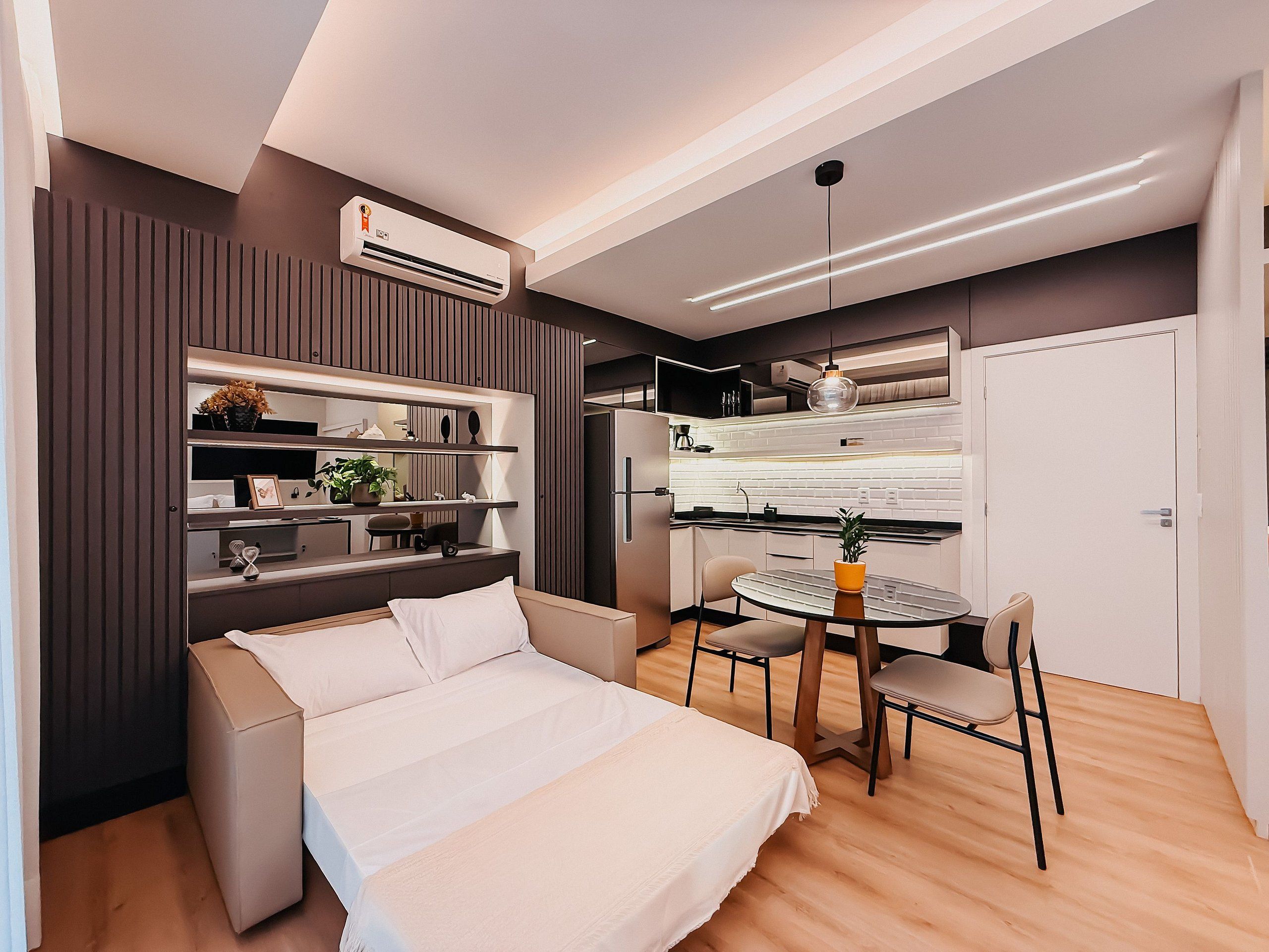 A modern bedroom with a bed, bedside tables, a small round table with chairs, a built-in wardrobe, and a kitchenette area.