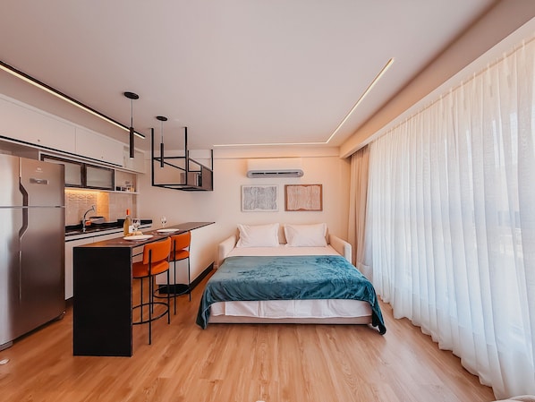 Una habitación de hotel compacta con kitchenette, una cama con colcha color turquesa y un ventanal con cortinas traslúcidas.
