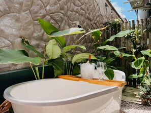 Una bañera con una bandeja de madera, una botella y dos vasos, rodeada de exuberante follaje verde.