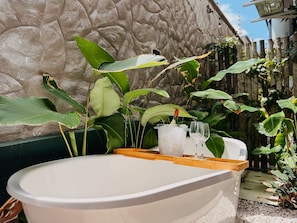 A bathtub with a wooden tray, a bottle, and two glasses, surrounded by lush green plants.