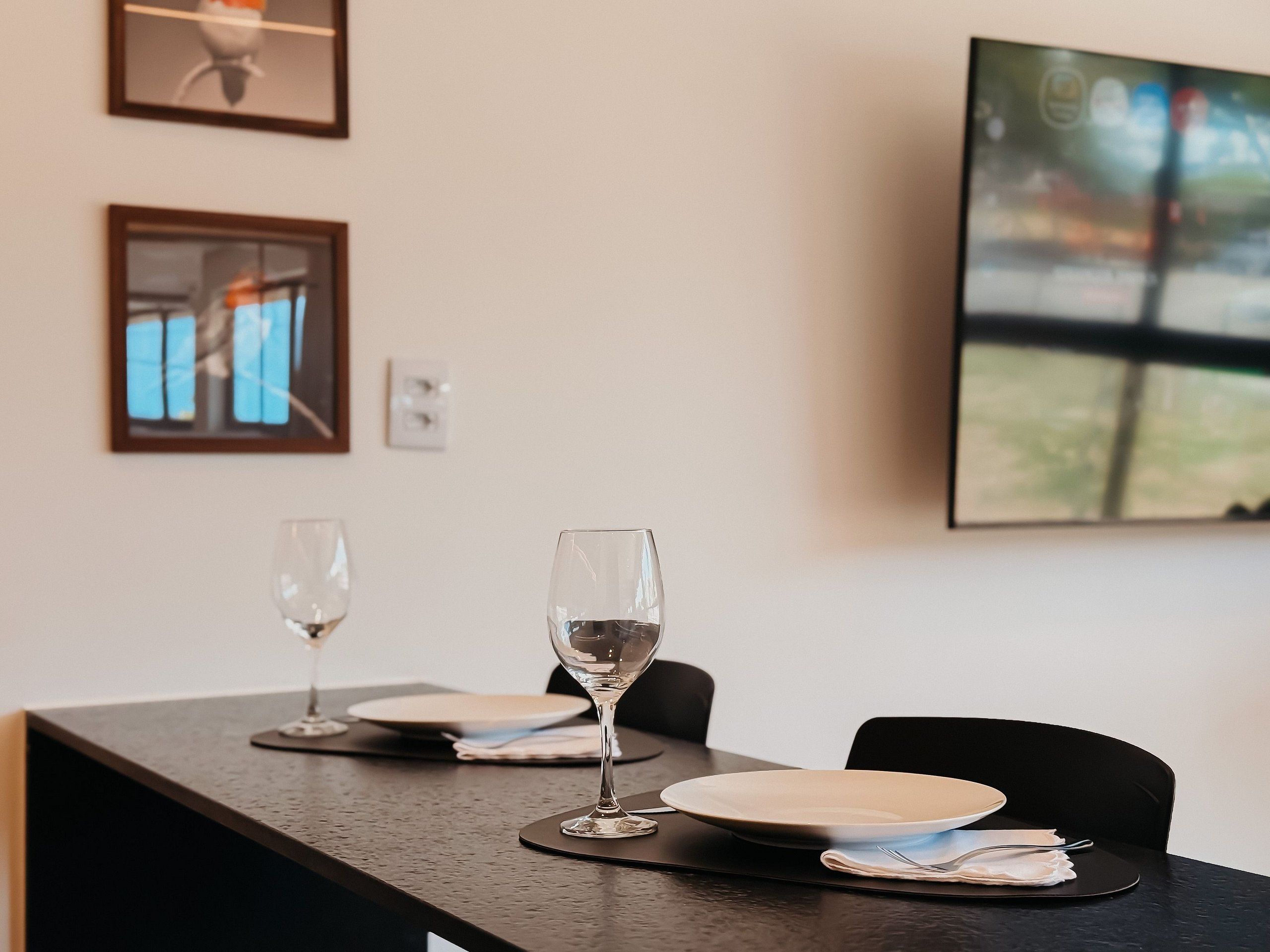 Une table à manger avec deux verres à vin, des assiettes et des couverts, placée devant un mur orné d’œuvres d’art encadrées et sur lequel est accrochée une télévision.