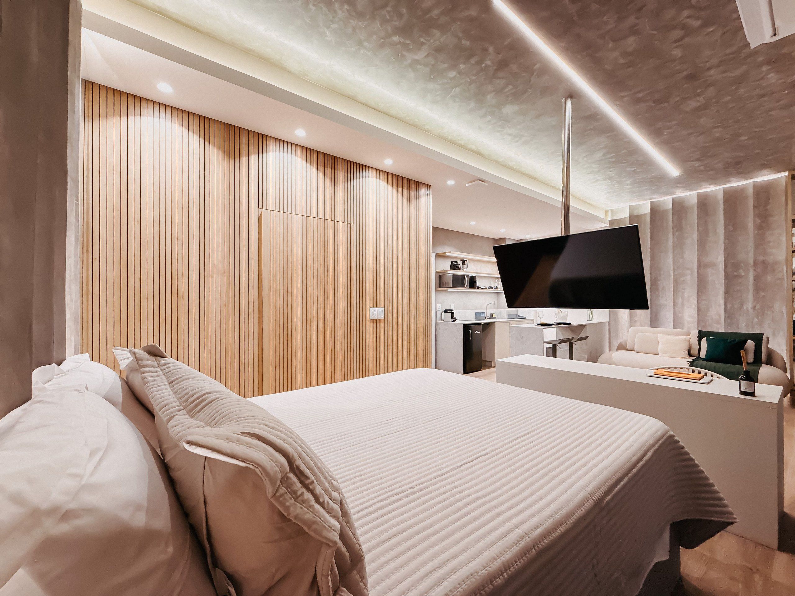Ett modernt hotellrum med en stor säng, en platt-TV som är monterad på väggen, ett litet kök och en sittgrupp med en soffa.