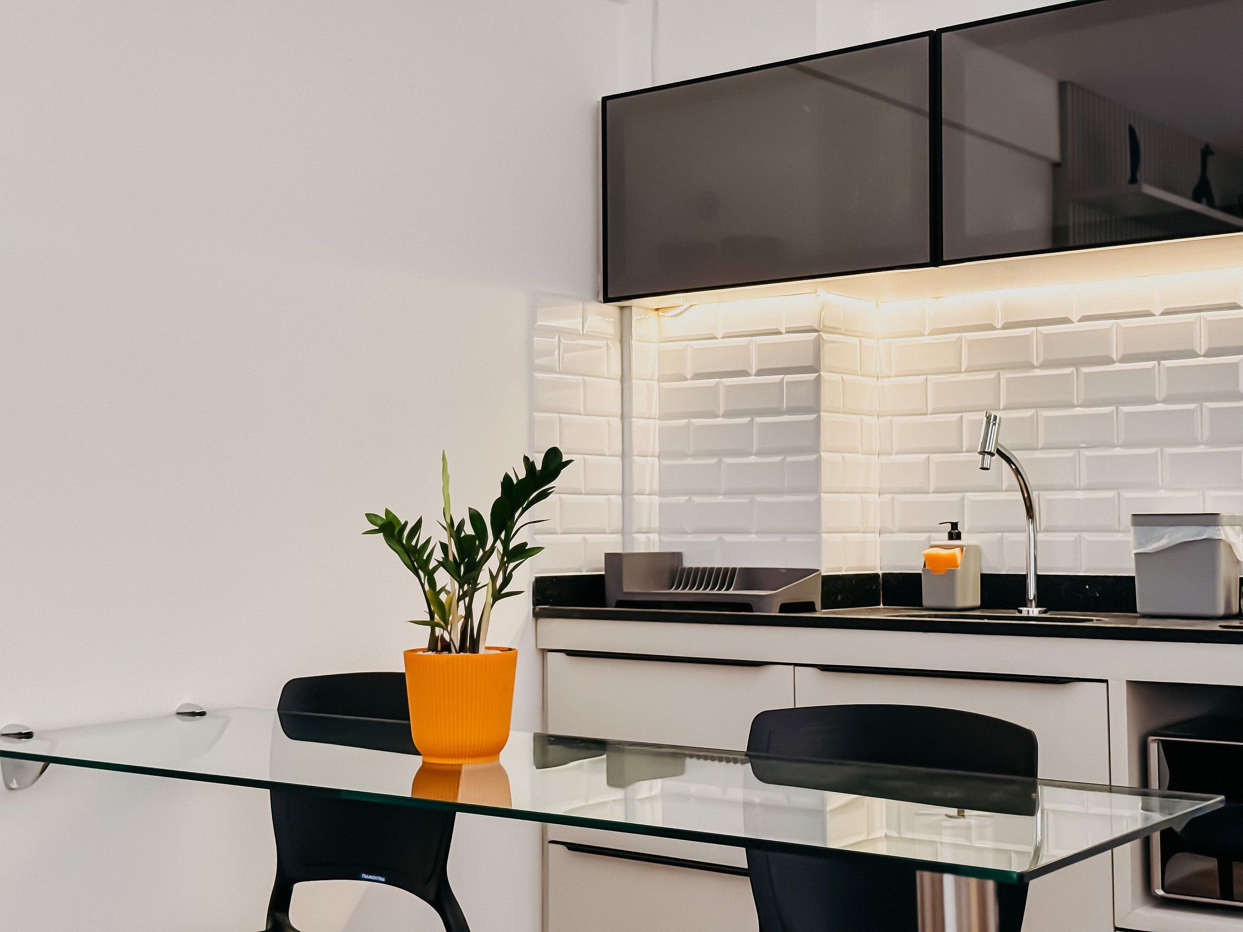Una cocina moderna con una mesa de vidrio, sillas negras, una planta en maceta, un fregadero y un televisor de pantalla plana montado sobre la encimera.