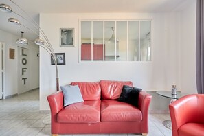 Un canapé en cuir rouge avec des coussins, une table basse en verre et une vue sur une cuisine à travers une fenêtre.