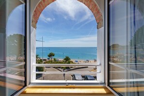 Blick durch ein Fenster mit Ziegelbogen auf einen Strand mit Menschen und Sonnenschirmen.