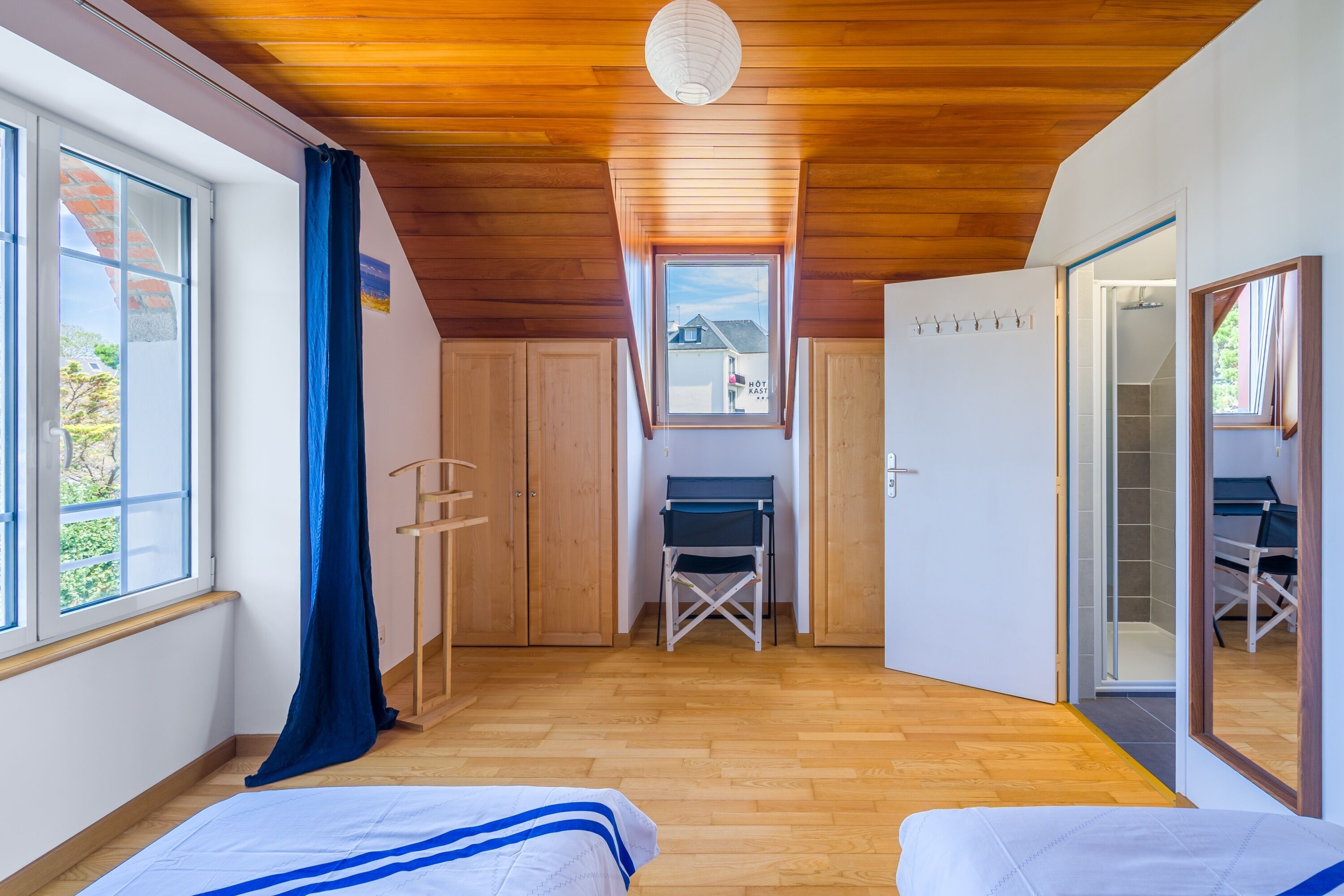 A room with a wooden ceiling, a window with a view of houses, a desk with a chair, and a bed with a blue-striped pillow.