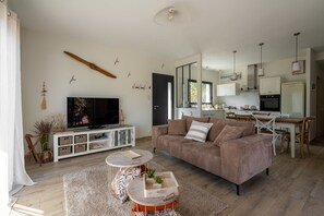 A modern living room with a large TV, a sofa, a coffee table, and a kitchen area in the background. - Les Hirondelles du Lac - Avec piscine privée (La Teste-de-Buch)