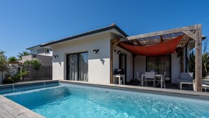 Una casa moderna con piscina, un patio cubierto con mobiliario y un jardín cercado.