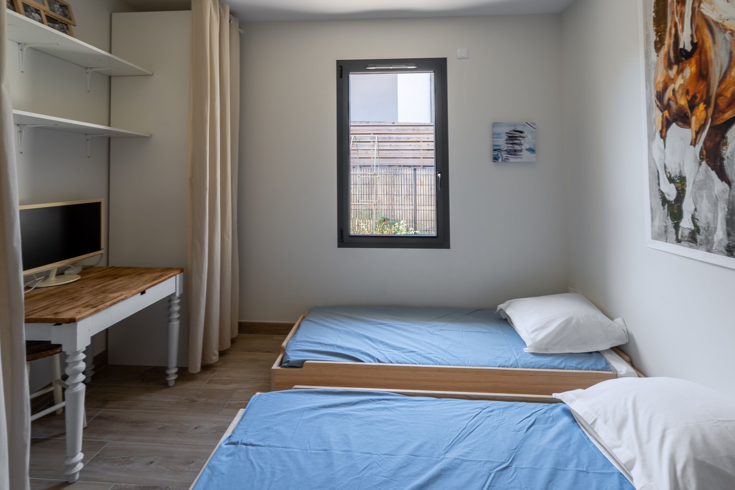 A bedroom with two single beds, a desk with a computer, a window, and a painting on the wall.