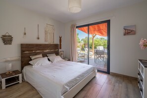 Ein Schlafzimmer mit einem großen Bett, einem Nachttisch, einer Wandlampe, einem Traumfänger, einem Holz-Kopfstück, einem gerahmten Bild und Blick ins Freie durch eine Schiebetür.