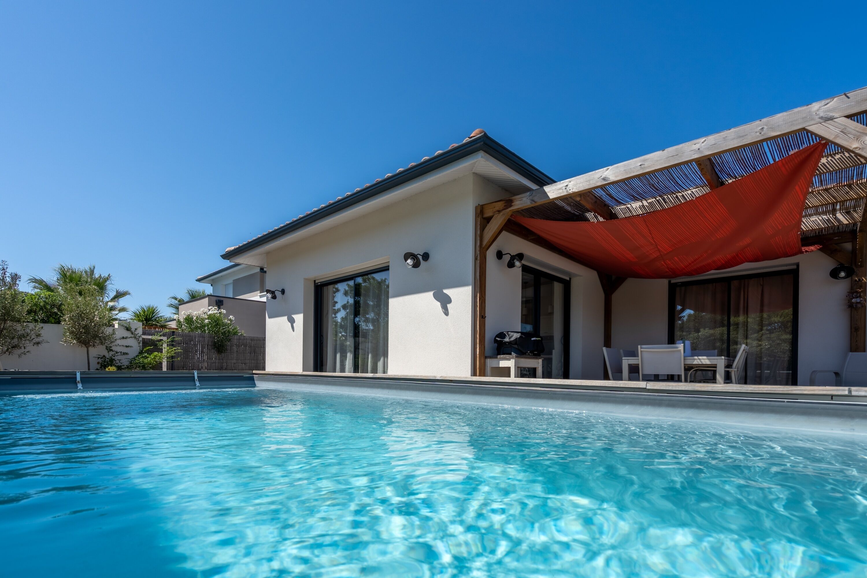 Une piscine à l’eau d’un bleu cristallin, un patio couvert aménagé avec du mobilier d’extérieur et un bâtiment moderne doté de grandes fenêtres.