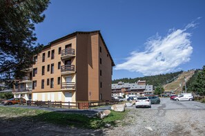 A multi-story building with balconies, a parking area with cars, and a hilly landscape in the background. - L’Écrin de Bolquère - Studio proche de la station (Bolquère)