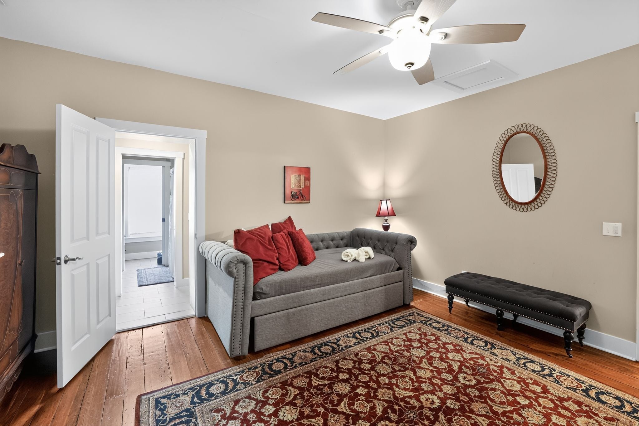 A living room with a grey sofa, a red throw blanket, a patterned rug, a bench, a ceiling fan, and a mirror on the wall.