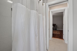 Um banheiro com cortina de chuveiro branca, parede revestida em azulejos e uma porta que dá para um quarto com tapete estampado e uma cama.