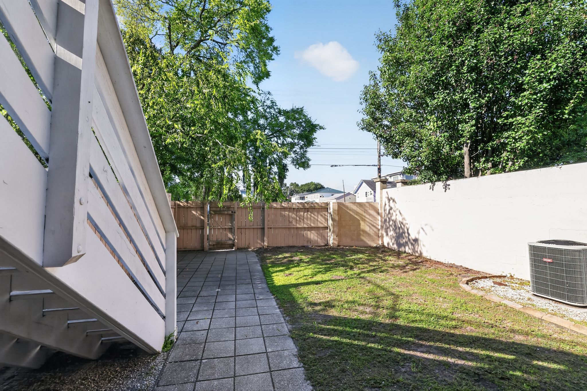 A paved pathway leading to a gated entrance, surrounded by a white wall and a fence, with trees and a clear sky in the background.