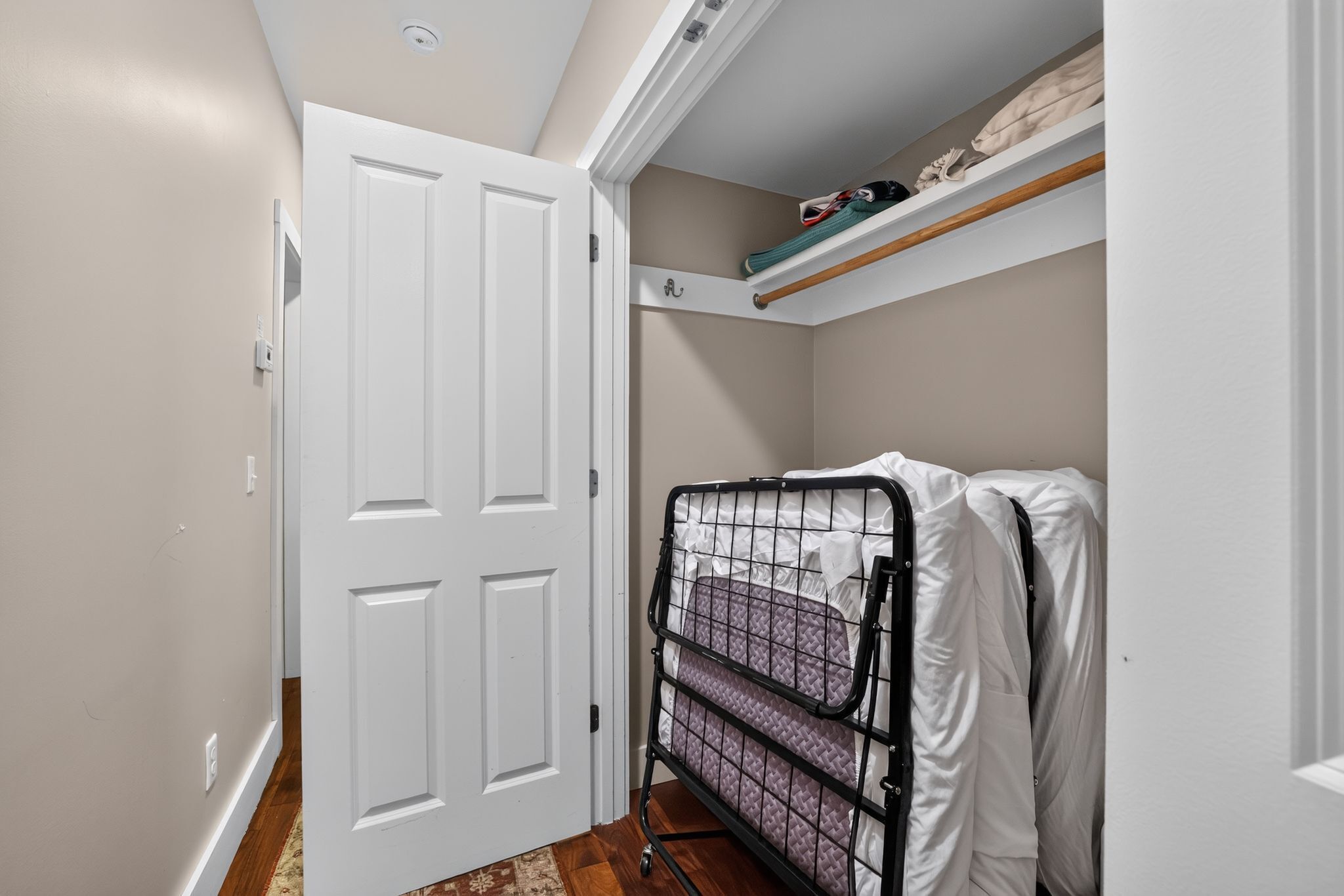 Un pasillo estrecho con una puerta blanca, una cama litera de metal con ropa de cama blanca y un estante sobre la cama.