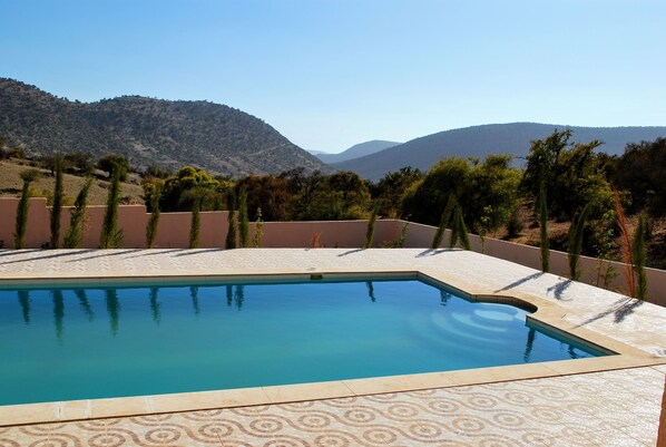A swimming pool with a patterned tile edge, surrounded by a landscaped area with trees and a view of mountains.