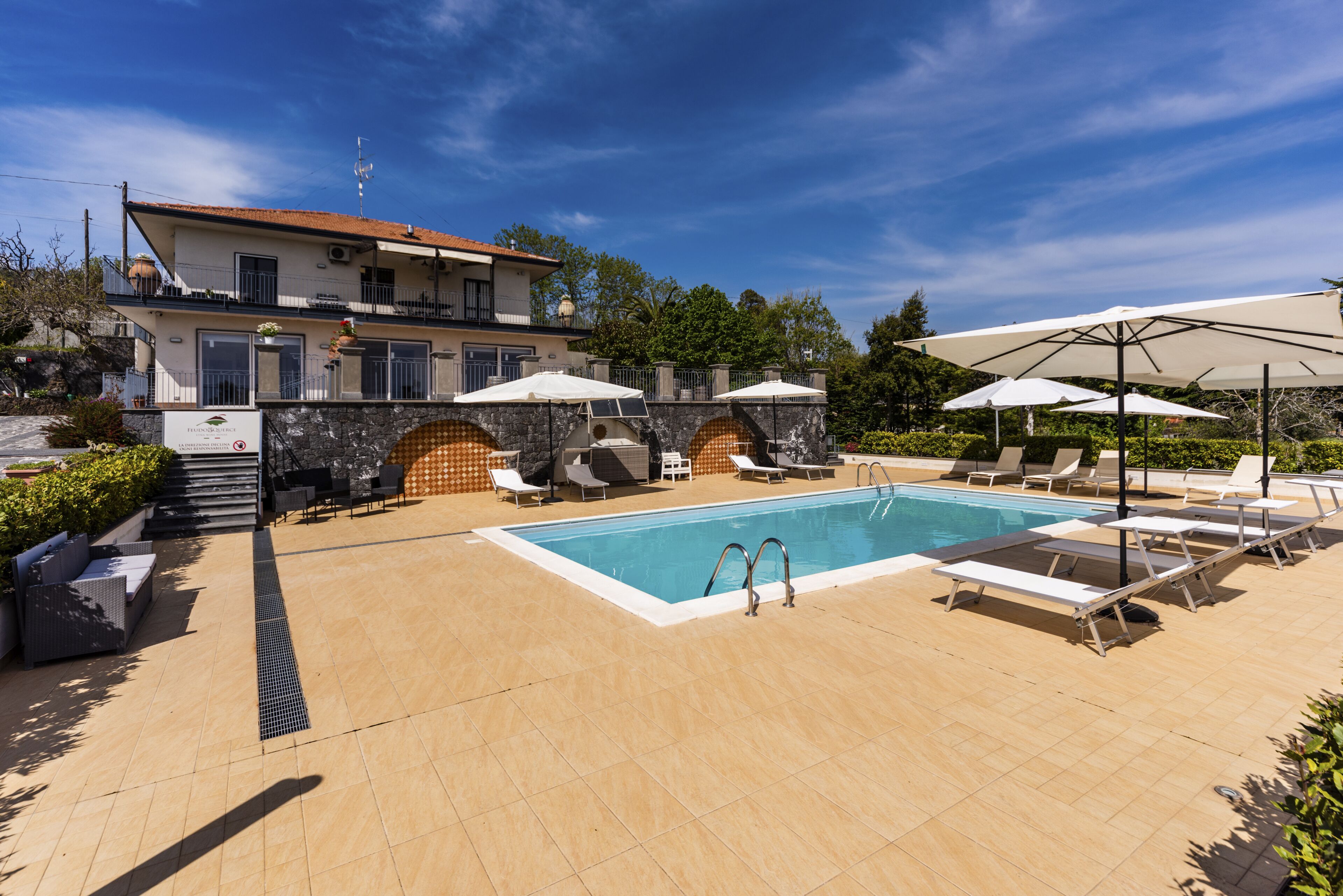 Zona de piscina con sillones reclinables y sombrillas, un muro de piedra y un edificio con balcón.