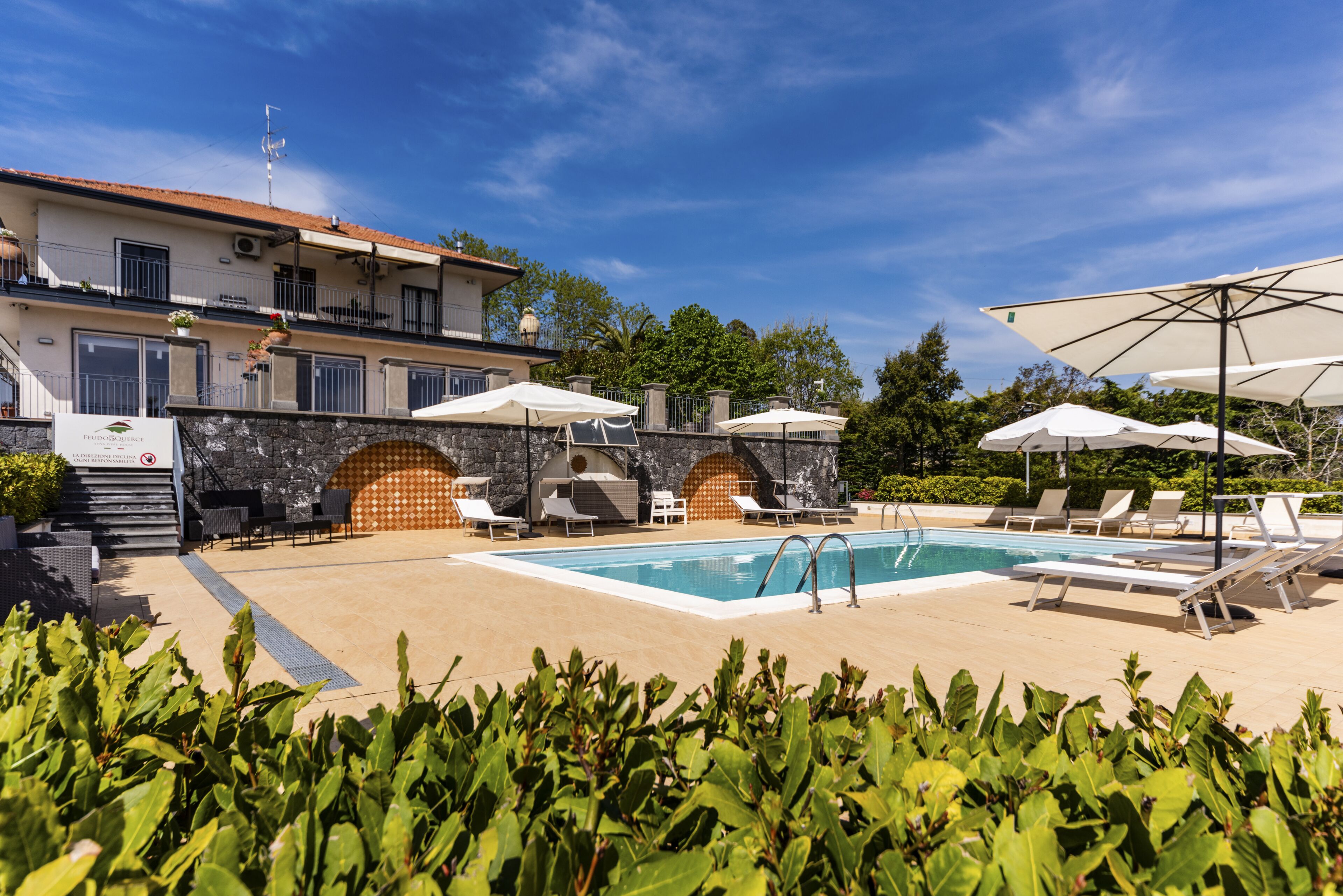 Zona de piscina con sillones de descanso y sombrillas, un muro de piedra y un edificio con techo de tejas.