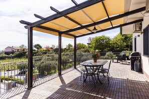 An outdoor patio with a table and chairs, a pergola with a canopy, and a view of a garden and houses in the background. - Villa Etna Pool & Seaview by Wonderful Italy (Belpasso)