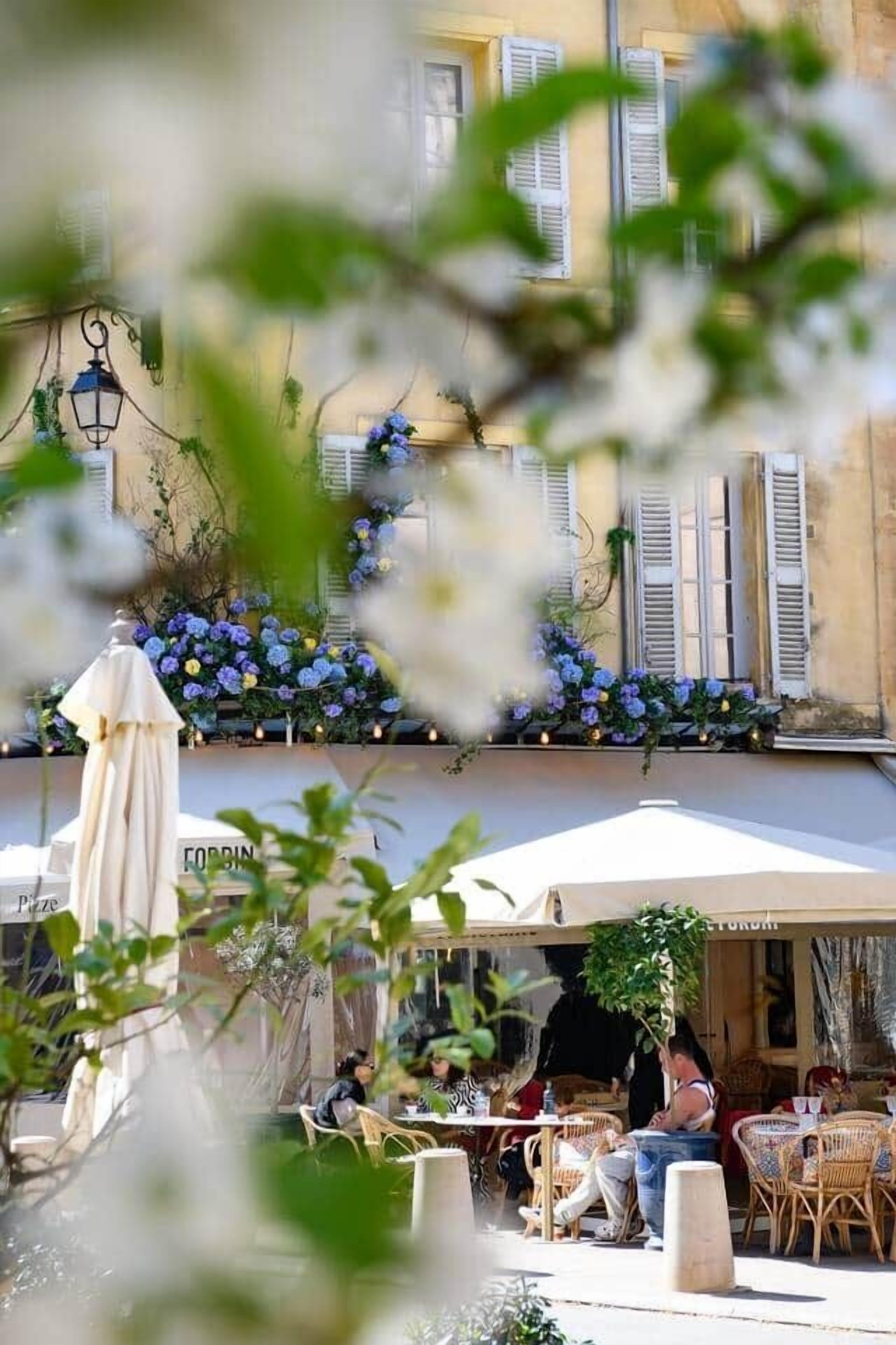 An outdoor cafe with tables and chairs, a white umbrella, and a building with shutters and flowers.
