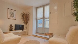 A living room with a sofa, a television, a decorative plant, and a window with curtains. - Les Contemporains-2 Side-by-Side Apartments – City Centre (Aix-en-Provence)