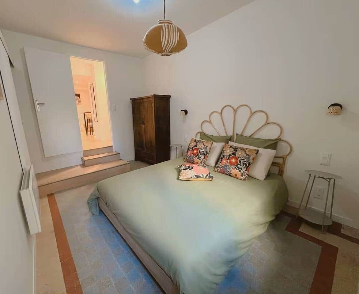 A bedroom with a bed, a wooden wardrobe, a small table, and a door leading to another room.