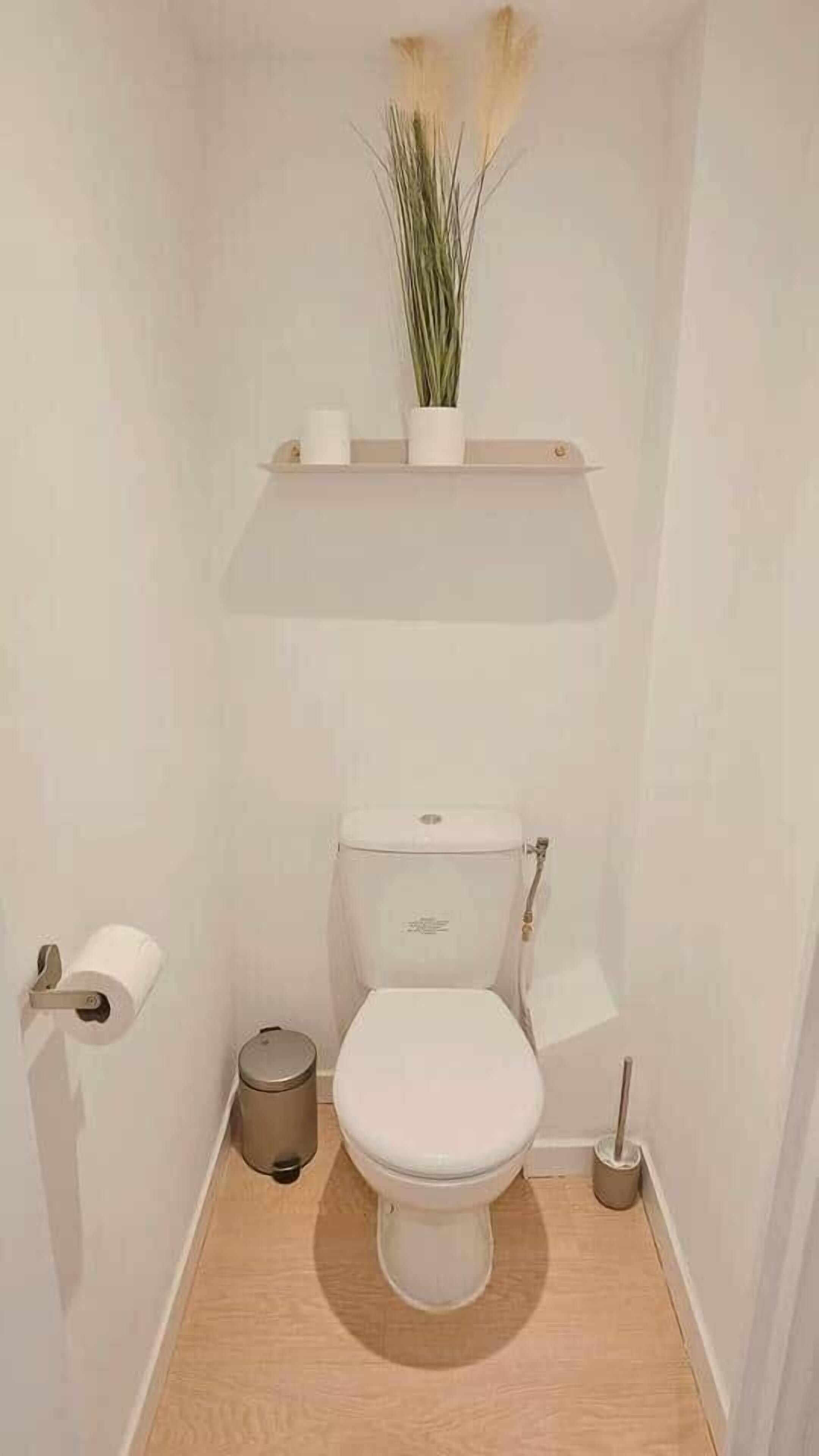 A small, modern bathroom with a toilet, a trash bin, a toilet paper holder, and a plant on a shelf.