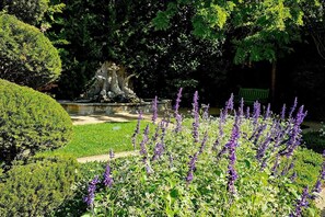 Ein gepflegter Garten mit Springbrunnen, geschnittenen Hecken und violetten Blumen.