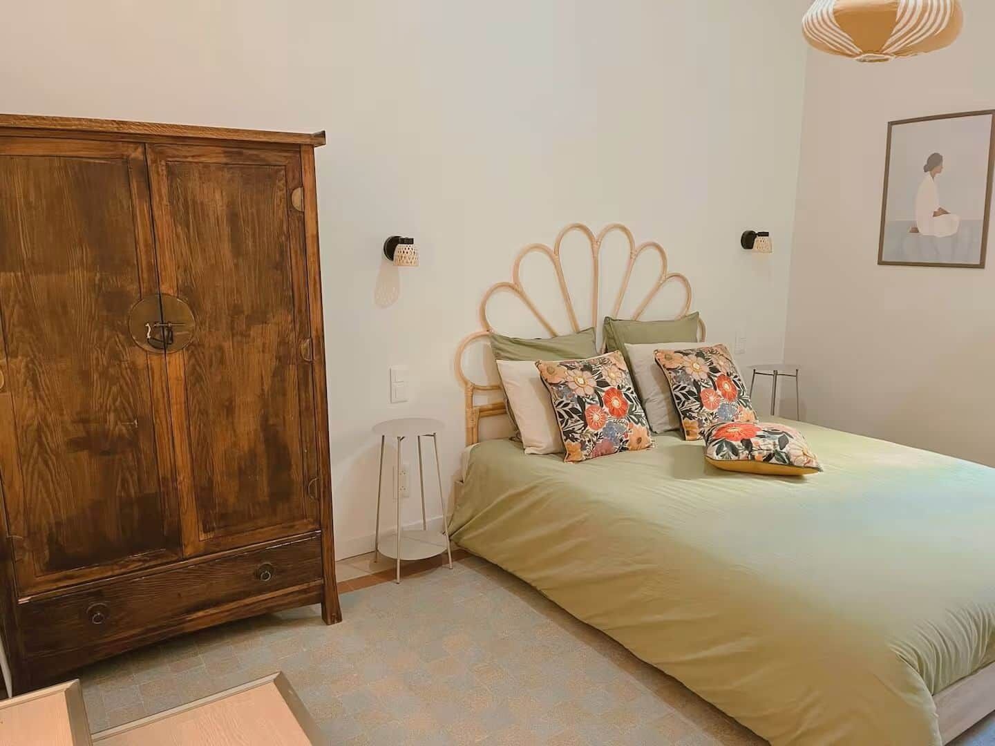 Une chambre avec une armoire en bois, un lit agrémenté d’oreillers à motifs floraux et un luminaire fixé au mur.