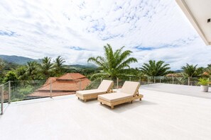 A balcony with two lounge chairs, a glass railing, and a view of a red-roofed building and palm trees. - Exclusive Golf Villa at Plam Hill  (Cha-am)