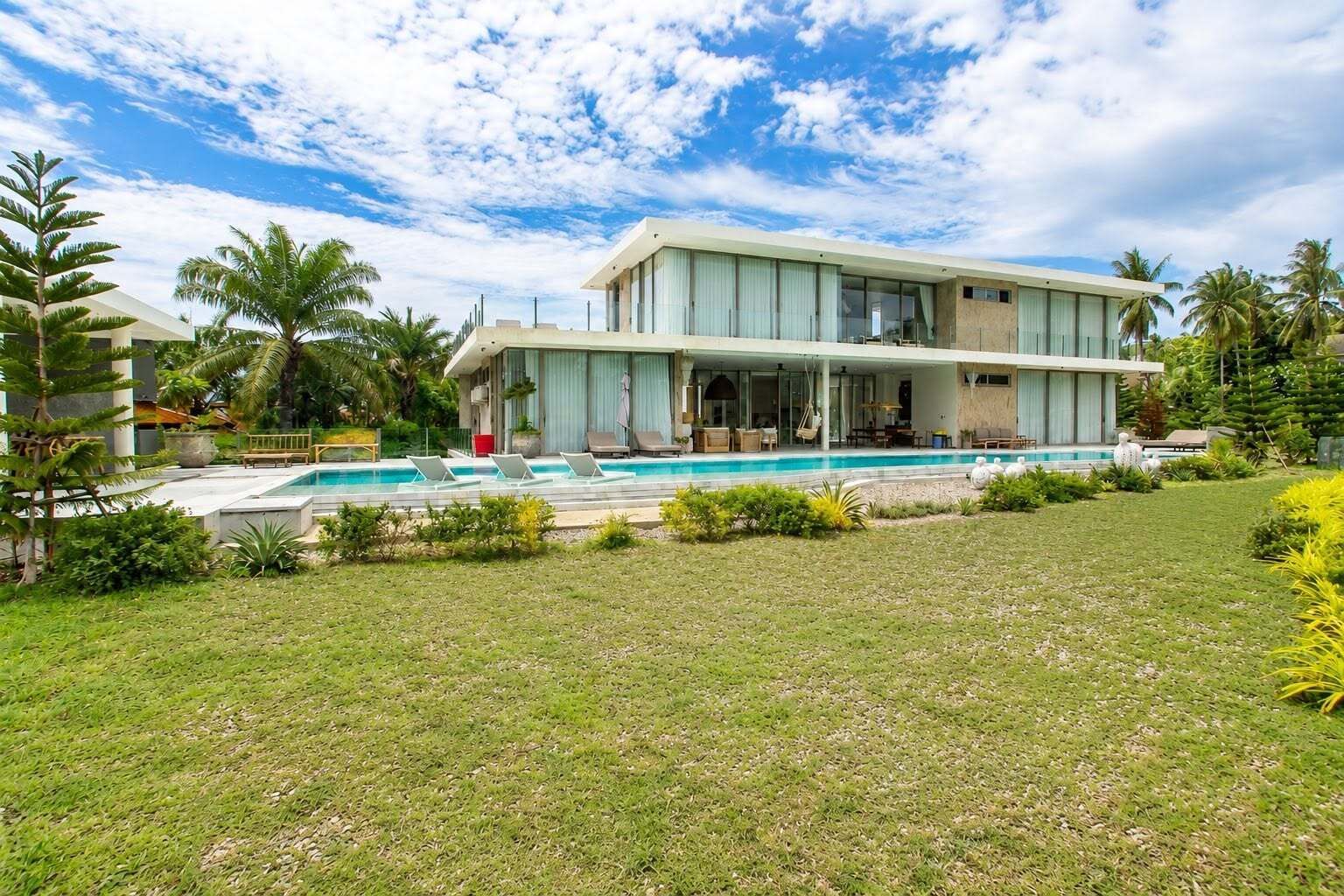 Una casa moderna de dos pisos con piscina, rodeada de exuberante vegetación y palmeras.