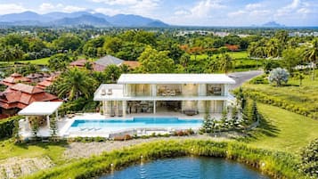 A modern two-story house with a swimming pool, surrounded by lush greenery and a view of distant mountains.