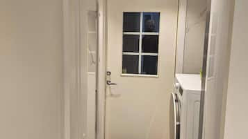 Un pasillo estrecho con una puerta, un ventanal pequeño, una lavadora y un refrigerador.