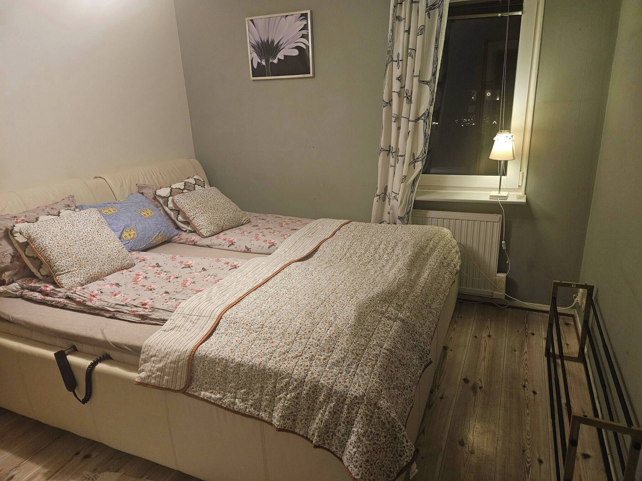 A single bed with floral bedding and multiple pillows, a bedside table with a lamp, a radiator, and a window with curtains.