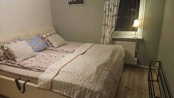 A single bed with floral bedding and multiple pillows, a bedside table with a lamp, a radiator, and a window with curtains.