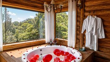 A wooden cabin with a large window, a hot tub with red balloons, and white bathrobes on the wall.