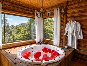 A wooden cabin with a large window, a hot tub with red balloons, and white bathrobes on the wall.