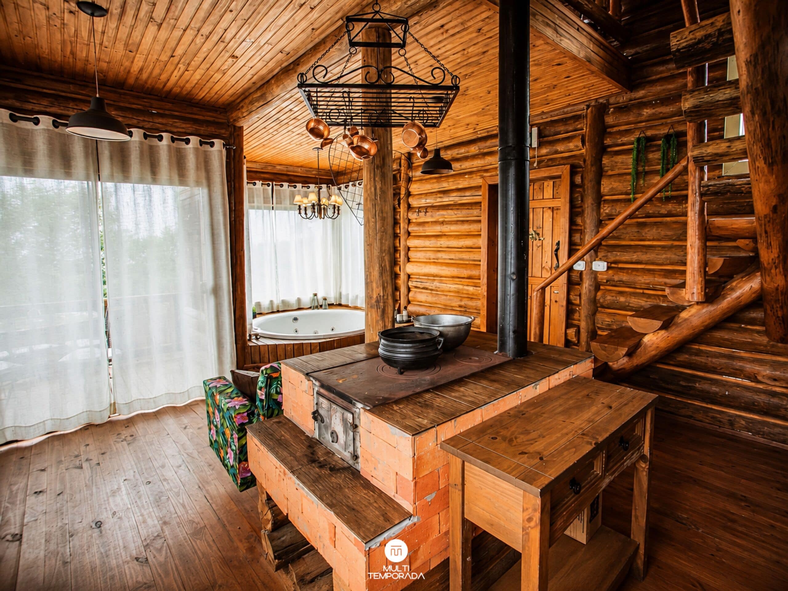 Interior rústico de una cabaña con detalles de madera, un ventanal con cortinas, un banco de madera, una mesa con un hervidor y una chimenea.